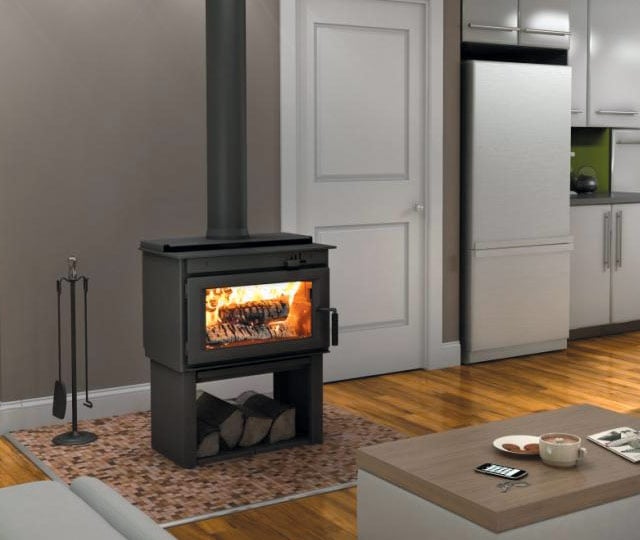 Modern living room with a wood-burning stove radiating warmth, placed on a tiled hearth mat. The stove has logs burning inside, with extra logs stored underneath. 