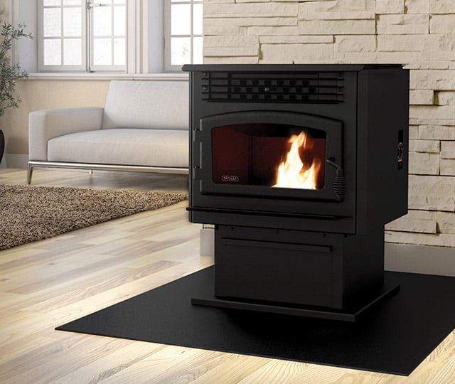 A sleek black pellet stove with a visible flame burns warmly in a modern, minimalist living room. The stove is positioned on a black mat over light wood flooring, near a beige textured wall and adjacent to a cozy white couch.